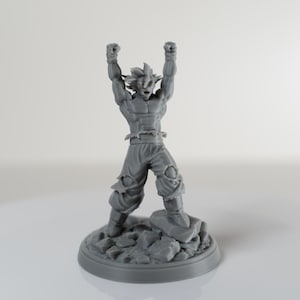 Może przedstawiać: Szara figurka muskularnego męskiego bohatera z kolczastymi włosami, z ramionami uniesionymi w triumfalnej pozie. Figurka stoi na skalistej podstawie. Figurka jest przedmiotem kolekcjonerskim.