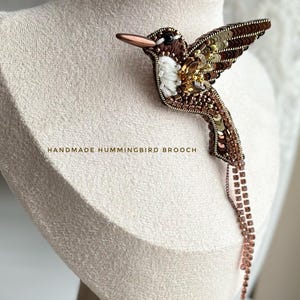 May include: A handmade hummingbird brooch with brown, gold, and white beadwork and sequin detailing. The brooch has a long, dangling chain of small, square-shaped beads. The text "HANDMADE HUMMINGBIRD BROOCH" is visible.