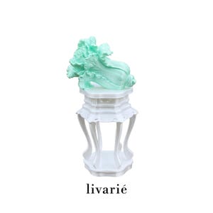 May include: A mint green cabbage-shaped sculpture resting on a white, multi-tiered pedestal stand with curved legs. The base of the stand features a scalloped edge design. The text "livarié" is printed in black below the white pedestal stand.