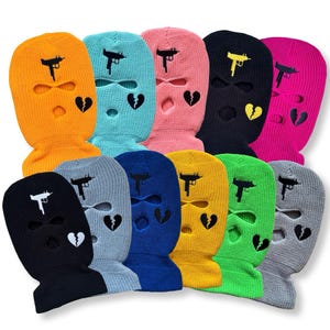 May include: A selection of knitted balaclavas in various colours. Each balaclava has a gun graphic and a broken heart design. Colours include orange, blue, pink, black, and yellow. The balaclavas have three holes for the eyes and mouth.