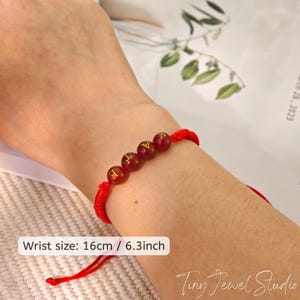 May include: Red bracelet featuring four reddish-brown beads with gold lettering. The bracelet is on a wrist, with the text "Wrist size: 16cm / 6.3inch" visible. The bracelet is made of red cord.
