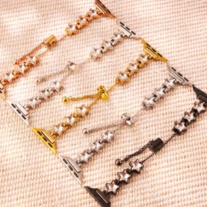 May include: An assortment of star-shaped watch bands in various metallic finishes, including gold, silver, and black. Each band features a chain with dangling star charms and a clasp for secure attachment to a smartwatch. The bands are displayed on a textured surface.