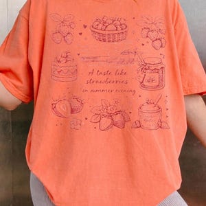 May include: Coral-colored t-shirt featuring a strawberry-themed design. The graphic includes illustrations of strawberries, a basket of berries, a jar of jam, and the text "A taste like strawberries in summer evening."
