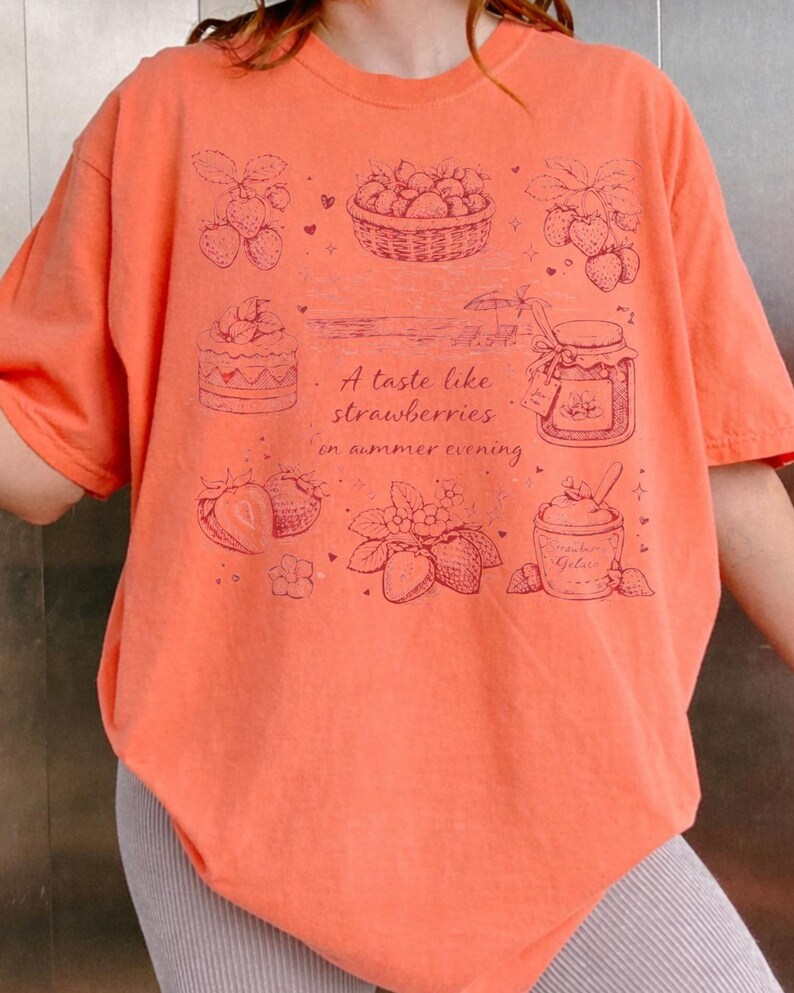 May include: Coral-colored t-shirt featuring a strawberry-themed design. The graphic includes illustrations of strawberries, a basket of berries, a jar of jam, and the text "A taste like strawberries in summer evening."