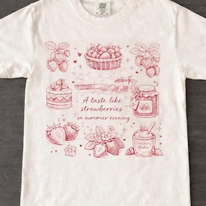 May include: White t-shirt with a red illustration of strawberries, a basket of berries, a cake, and a jar of jam. The text "A taste like strawberries on summer evening" is also printed on the shirt.