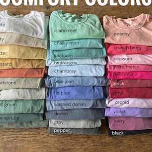 May include: Stacks of folded t-shirts in various colors, including white, yellow, orange, green, and blue. The text "COMFORT COLORS" is at the top. Each shirt is labeled with its color name, such as "island reef" and "peachy".
