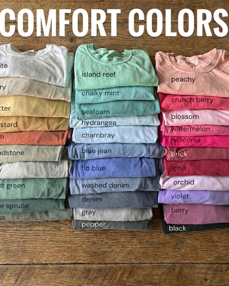 May include: Stacks of folded t-shirts in various colors, including white, yellow, orange, green, and blue. The text "COMFORT COLORS" is at the top. Each shirt is labeled with its color name, such as "island reef" and "peachy".