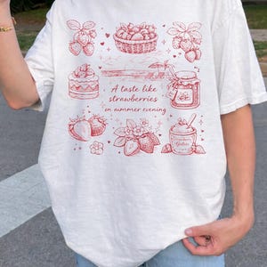 May include: White t-shirt with a red illustration of strawberries, a basket of strawberries, a jar of jam, and strawberry gelato. The text "A taste like strawberries on summer evening" is also printed on the shirt.