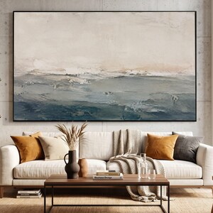 May include: A large rectangular abstract painting in a black frame featuring horizontal layers of cream, beige, and deep blue tones with visible textured brushstrokes, displayed above a white sofa with orange and grey throw pillows and a wooden table.