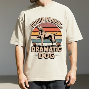 May include: Beige t-shirt with a graphic featuring a dog silhouette inside a sunset design. The text reads "PROUD PARENT OF DRAMATIC DOG" in a vintage style font. The shirt is a relaxed fit.