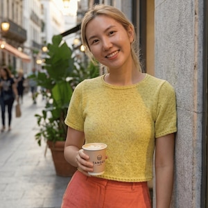 May include: A woman wearing a yellow short-sleeved knit top and orange shorts holds a coffee cup. The cup has the text "Tanger Cafe" printed on it. The background shows a European street scene with buildings and greenery.