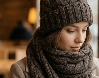 Puede incluir: Un conjunto de gorro y bufanda de punto trenzado marrón. El gorro tiene un borde doblado y un diseño tejido texturizado. La bufanda está envuelta alrededor del cuello y los hombros. El conjunto está hecho de un material suave y cálido.