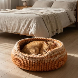 May include: An orange tabby cat sleeps peacefully in a round, hand-knitted cat bed. The bed is made from orange and cream-coloured yarn, with a chunky, textured design. The cat is curled up inside the bed. The bed is on a wooden floor.