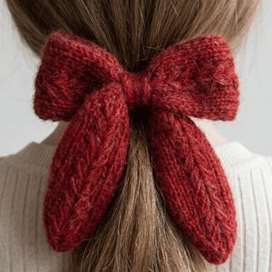Op de afbeelding: Een close-up van een rode gebreide strik haaraccessoire. De strik is gemaakt van een getextureerd garen en is in een klassieke strikvorm geknoopt. Het haaraccessoire wordt in het haar gedragen, met de strik aan de achterkant van het hoofd.