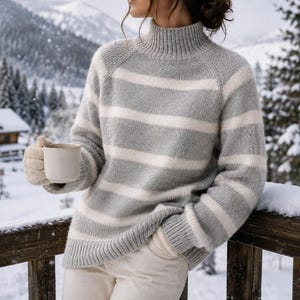 Op de afbeelding: Een gebreide trui met lange mouwen, een hoge col en horizontale strepen in afwisselende tinten lichtgrijs en off-white. Het kledingstuk heeft geribbelde boorden aan de mouwen en de onderkant, gecombineerd met een lichte broek en textuurwanten.