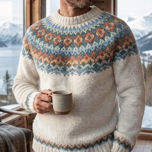 Può includere: Un maglione a maniche lunghe color crema con scollo rotondo e orlo a coste. Il capo presenta un dettagliato motivo geometrico sul petto e sui polsini in tonalità blu, arancione e marrone. Una tazza in ceramica è tenuta in una mano.