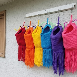 May include: A row of six colorful, knitted monster-like pouches hanging on a white rack. Each pouch is a different color: red, orange, yellow, blue, purple, and pink. They have googly eyes, antennae, and fringe.