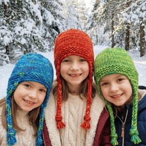 Könnte beinhalten: Drei Kinder tragen bunte gestrickte Wintermützen mit geflochtenen Bändern. Die Mützen sind in Blau, Rot und Grün gehalten. Der Hintergrund ist eine verschneite Waldszene.
