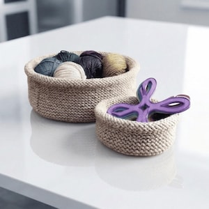Pode incluir: Duas cestas de malha bege. A cesta maior contém várias bolas de fio em várias cores, incluindo azul, bege e roxo. A cesta menor contém um objeto decorativo roxo multicamadas. As cestas estão em uma superfície branca.