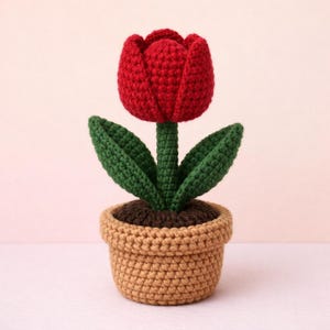 Może przedstawiać: Ręcznie robiony szydełkowy tulipan w doniczce. Czerwony tulipan ma zielone liście i brązową łodygę. Kwiat znajduje się w beżowej doniczce z brązową „ziemią”. Szydełkowanie jest szczegółowe, co czyni go uroczym przedmiotem dekoracyjnym.
