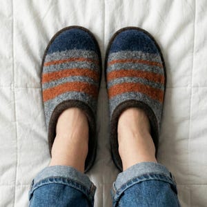 Op de afbeelding: Een paar gestreepte wollen pantoffels in blauwe, grijze en oranje tinten. De pantoffels hebben een donkerbruine rand en worden getoond op een wit gewatteerd oppervlak. De persoon draagt een blauwe spijkerbroek.