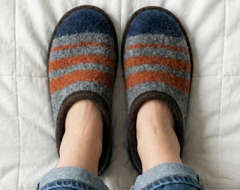 Puede incluir: Un par de zapatillas de lana a rayas en tonos azules, grises y naranjas. Las zapatillas tienen un ribete marrón oscuro y se muestran sobre una superficie acolchada blanca. La persona lleva vaqueros azules.