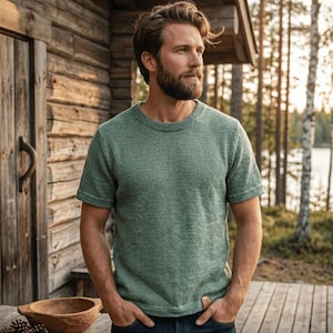 Puede incluir: Una camiseta de punto verde salvia de manga corta con cuello redondo. La camiseta tiene un aspecto texturizado y una peque&ntilde;a etiqueta rectangular en la parte inferior derecha. La persona lleva vaqueros azules y est&aacute; de pie frente a una caba&ntilde;a de madera.