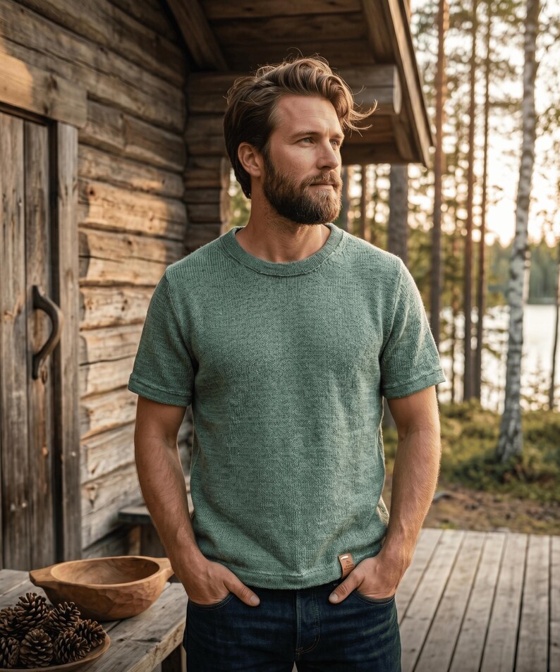 Puede incluir: Una camiseta de punto verde salvia de manga corta con cuello redondo. La camiseta tiene un aspecto texturizado y una peque&ntilde;a etiqueta rectangular en la parte inferior derecha. La persona lleva vaqueros azules y est&aacute; de pie frente a una caba&ntilde;a de madera.