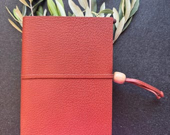 May include: A red leather-bound journal with a textured cover, secured by an elastic band and a wooden bead. A tassel hangs from the bottom. Olive branches with green and light green leaves are arranged behind the journal against a dark background.