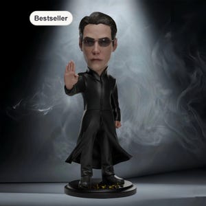 Puede incluir: Figura de bobblehead de un hombre con abrigo negro largo y gafas de sol, con la mano derecha levantada. La figura está sobre una base negra con detalles dorados. La palabra "Bestseller" está en un óvalo blanco.