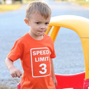 May include: A young boy wearing an orange t-shirt with a white "Speed Limit 3" sign graphic. The boy is standing in front of a yellow and red play structure.