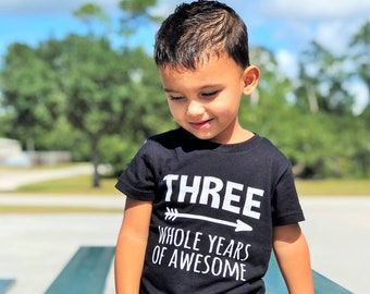Może przedstawiać: Małe dziecko w czarnym t-shircie z białym napisem "THREE WHOLE YEARS OF AWESOME". Dziecko ma na sobie podarte dżinsowe szorty. Tło przedstawia park z błękitnym niebem i drzewami.