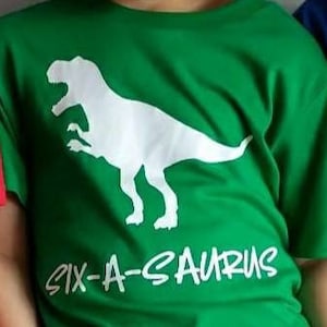 May include: A young boy wearing a green t-shirt with a white dinosaur graphic and the text "Six-A-Saurs".