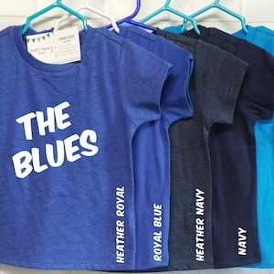 以下が含まれることがあります： ハンガーに掛けられた、さまざまな青色の5枚のTシャツ。Tシャツには、ヘザーロイヤル、ロイヤルブルー、ヘザーネイビー、ネイビー、アイランドブルーの色がラベル付けされています。一番前のTシャツには、「THE BLUES」という白い文字が書かれています。
