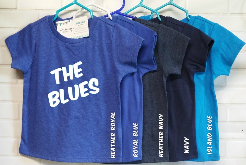 以下が含まれることがあります： ハンガーに掛けられた、さまざまな青色の5枚のTシャツ。Tシャツには、ヘザーロイヤル、ロイヤルブルー、ヘザーネイビー、ネイビー、アイランドブルーの色がラベル付けされています。一番前のTシャツには、「THE BLUES」という白い文字が書かれています。