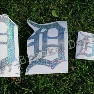May include: Four different sizes of silver glitter vinyl decals of the letter "D" in a gothic font. The decals are on a white background and are arranged in a row from largest to smallest.
