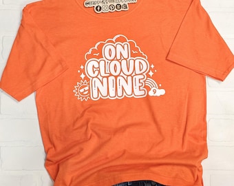 May include: Orange t-shirt with the phrase "ON CLOUD NINE" in white, stylized text. The design includes a cloud, rainbow, sun, and stars. The shirt is folded over blue jeans.