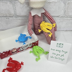 May include: A Christmas-themed toy set featuring a red and white striped elf doll, a white box with a Santa Claus design, and four colorful plastic frog toys. A card asks, "How many frogs can you get into the tray?"