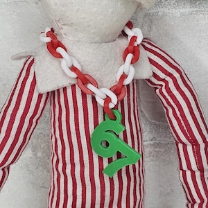 May include: A doll in a red and white striped outfit with a white collar. It is wearing a necklace of red and white chain links and a green number 67 pendant. The doll's outfit features vertical red and white stripes.