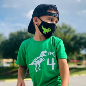 以下が含まれることがあります： 緑のTシャツを着た小さな子供が、白い恐竜のグラフィックと「Roar! I'm 4」の文字をプリントしています。子供は、緑の恐竜のグラフィックがプリントされた黒いフェイスマスクと、黒いベースボールキャップも着用しています。