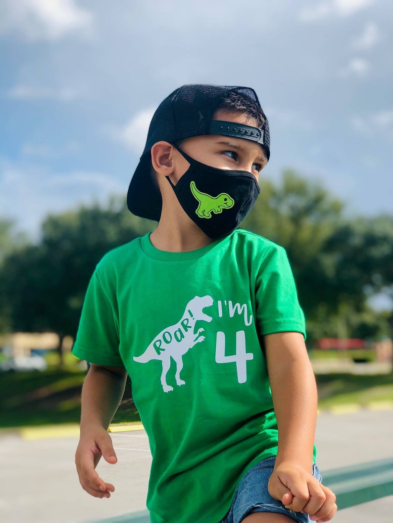以下が含まれることがあります： 緑のTシャツを着た小さな子供が、白い恐竜のグラフィックと「Roar! I'm 4」の文字をプリントしています。子供は、緑の恐竜のグラフィックがプリントされた黒いフェイスマスクと、黒いベースボールキャップも着用しています。