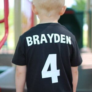 以下が含まれることがあります： 後ろに「Brayden 4」と白文字で書かれた黒いTシャツです。