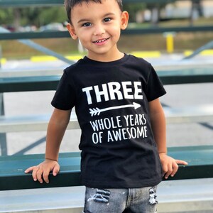 May include: A young child wearing a black t-shirt with the text "THREE WHOLE YEARS OF AWESOME" in white. The child is also wearing distressed denim jeans. The child is smiling and leaning on a green bench.