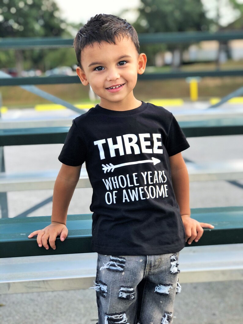 May include: A young child wearing a black t-shirt with the text "THREE WHOLE YEARS OF AWESOME" in white. The child is also wearing distressed denim jeans. The child is smiling and leaning on a green bench.