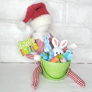 May include: A red and white striped elf doll holding a green basket filled with Easter-themed toys, including a white bunny, blue birds, and carrots. The elf is holding a sign that says "Happy Easter".