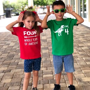 以下が含まれることがあります： 誕生日Tシャツを着た子供2人。女の子は「FOUR WHOLE YEARS OF AWESOME」と書かれた赤いTシャツを着ています。男の子は恐竜のグラフィックと「ROAR! I'M 4」と書かれた緑のTシャツを着ています。