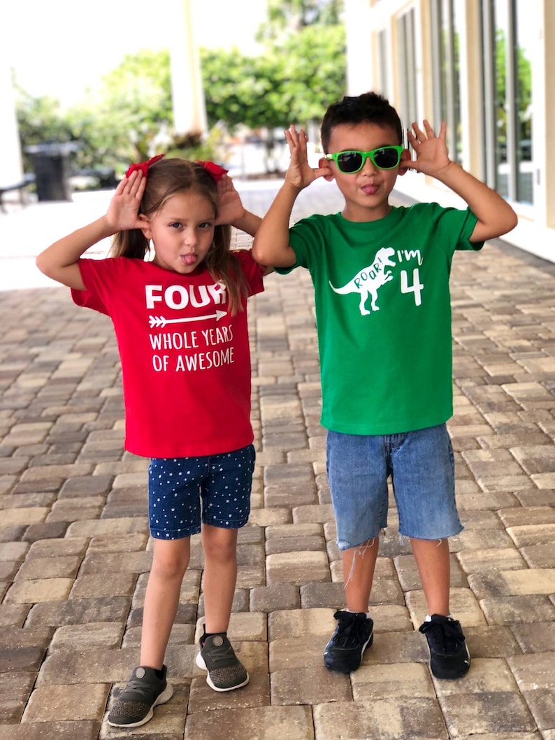 以下が含まれることがあります： 誕生日Tシャツを着た子供2人。女の子は「FOUR WHOLE YEARS OF AWESOME」と書かれた赤いTシャツを着ています。男の子は恐竜のグラフィックと「ROAR! I'M 4」と書かれた緑のTシャツを着ています。