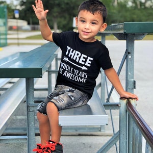 May include: A young child wearing a black t-shirt with the text "THREE WHOLE YEARS OF AWESOME" and distressed denim shorts. The child is sitting on a metal bench and wearing black and orange sneakers, waving at the camera.