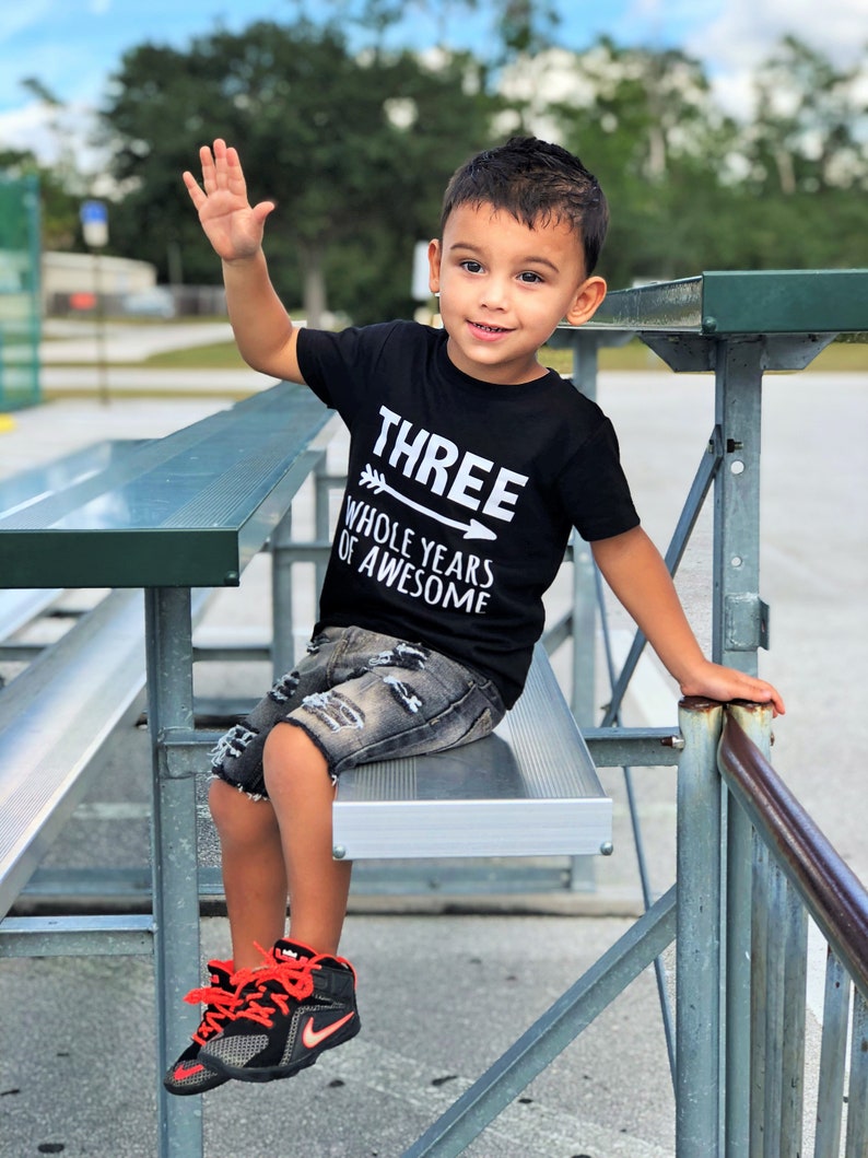 May include: A young child wearing a black t-shirt with the text "THREE WHOLE YEARS OF AWESOME" and distressed denim shorts. The child is sitting on a metal bench and wearing black and orange sneakers, waving at the camera.