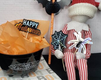 May include: A black cauldron with the text "Happy Halloween" on it, surrounded by orange tissue paper. A red and white striped elf doll is holding a black spider web and a silver spider with a black body. The elf is wearing a red knit hat with white trim.